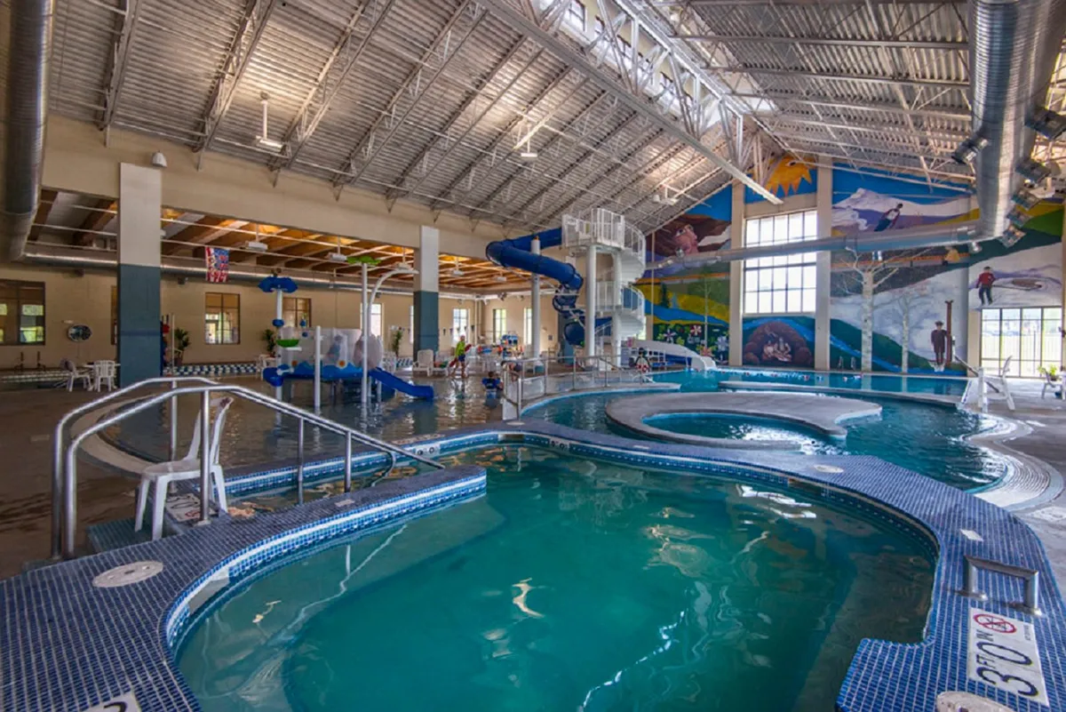 A vibrant indoor swimming pool area featuring a winding waterslide, colorful murals, and abundant lounge chairs. Children splash and play while families gather by the pool, creating an energetic and joyful atmosphere.