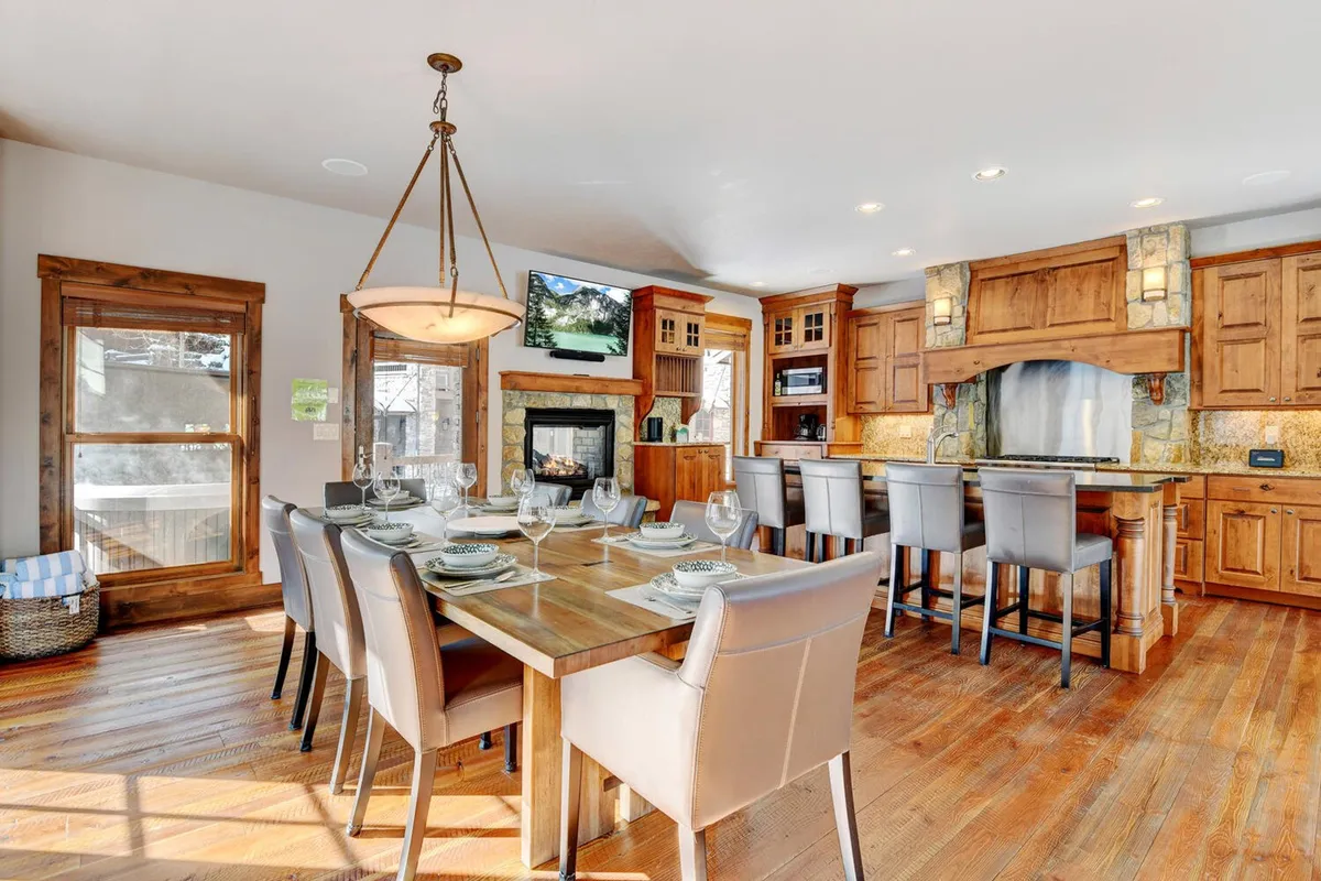 This interior image presents a spacious dining area with a table, kitchen island, fireplace, and large windows. The mood is warm and inviting, ideal for family dinners, entertaining guests, or relaxing by the fire, with mountain views visible through the windows.