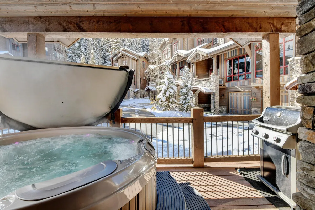 The private deck area with a snow-surrounded hot tub and a grill, offering an outdoor extension of the kitchen experience. The wooden deck sets a tranquil mood, perfect for grilling, enjoying a snack after a day of adventure, or relaxing in the hot tub under the stars. The scene connects indoor culinary experiences with the outdoors.
