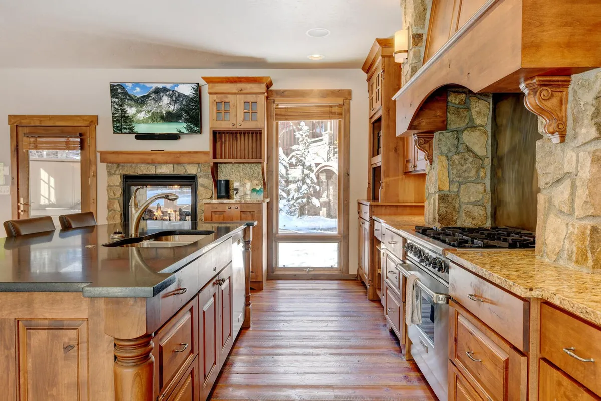 A spacious, cozy kitchen featuring a large island with stools, a striking stone fireplace, wooden cabinetry, stainless steel appliances, and a large window letting in natural light. The kitchen island is set up for both cooking and gathering, while the fireplace hints at warmth and ambiance. All elements feel both functional and inviting, perfectly suited for both elaborate meals and casual breakfasts.