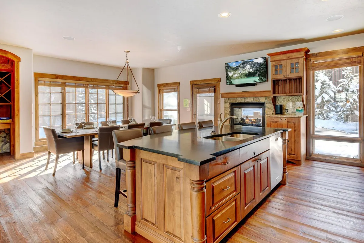An elegant open-plan space blends the kitchen, dining area, and living room. A stone fireplace, large windows overlooking snowy landscapes, and modern furnishings create a seamless, welcoming environment for groups to gather and unwind.