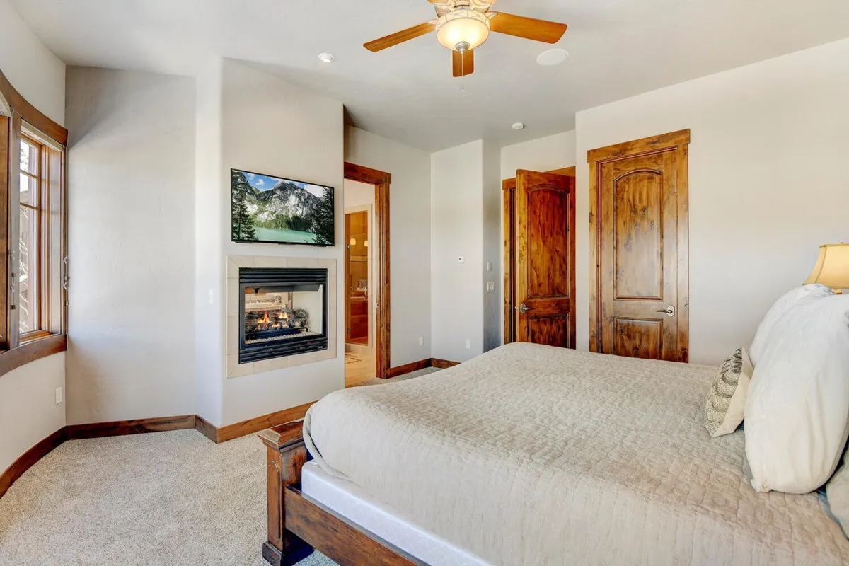 A spacious king bedroom featuring a plush bed, elegant fireplace, inviting window seating, and tasteful wall art. The glow from the double-sided fireplace creates a serene atmosphere that extends into the adjoining areas, suggesting both restful luxury and a unique connection between bedroom and bath.
