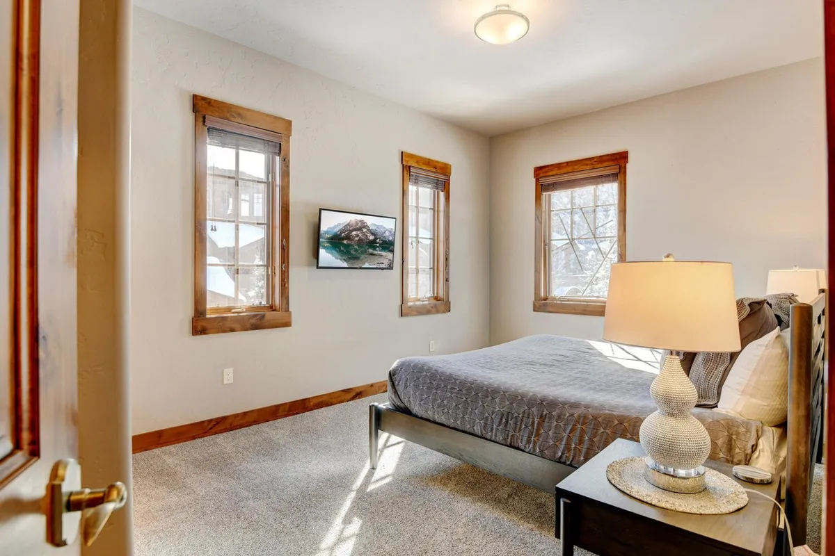 A cozy adult bedroom featuring a comfortable bed, bedside lamp, smart HDTV, and windows that let in natural light. The setting is perfect for unwinding with a favorite show or enjoying a peaceful morning.