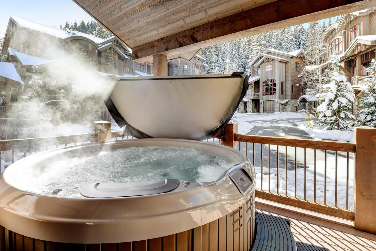 An exterior view of a wooden deck featuring a private hot tub surrounded by snow-covered trees. The scene exudes tranquility, with inviting steam rising from the tub and comfortable seating for relaxing. The mountains can be glimpsed in the distance, suggesting a direct connection to the outdoor adventures described.