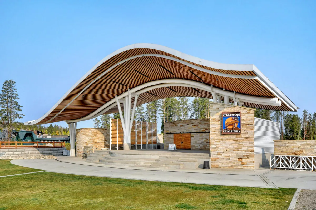 An exterior shot of an outdoor amphitheater with a distinctive curved roof, stone facade, and event signage. The space is set up for community events, concerts, and festivals, set against a scenic backdrop.