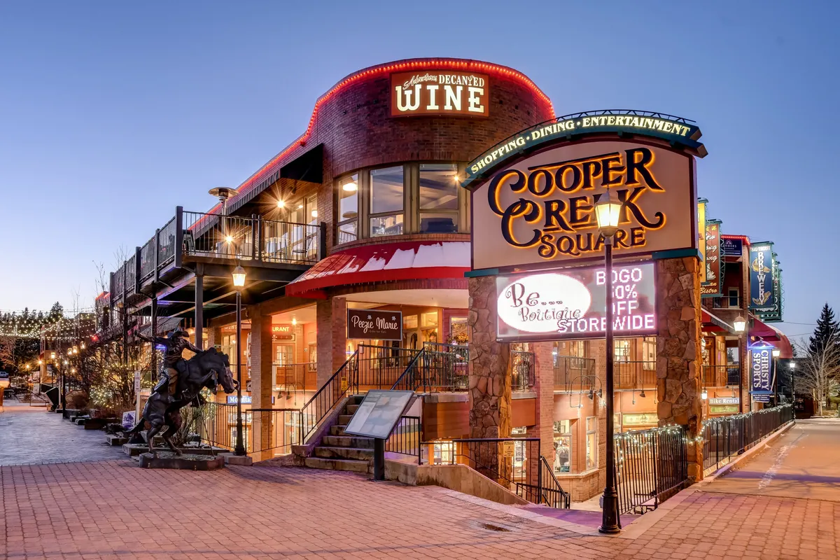 A bustling outdoor plaza scene at Cooper Creek Square, with visible signage, a wine shop, outdoor seating, and cheerful decorative lighting, hinting at shopping and dining opportunities.