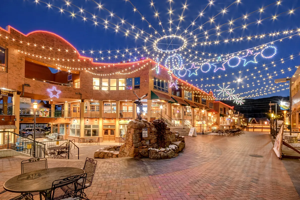 A lively exterior plaza adorned with string lights and decorative stars, featuring a stone fountain and inviting outdoor seating. The scene suggests an atmosphere of celebration, community, and leisure.