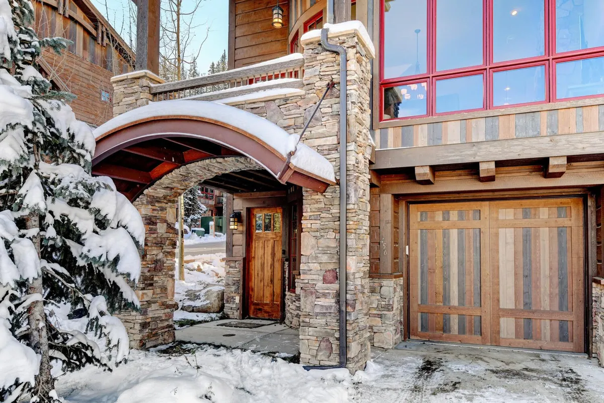 This image features the arched stone entryway of the property, a wooden front door flanked by large red windows, all set in a snow-covered landscape. The inviting exterior hints at both architectural elegance and the allure of a winter retreat.